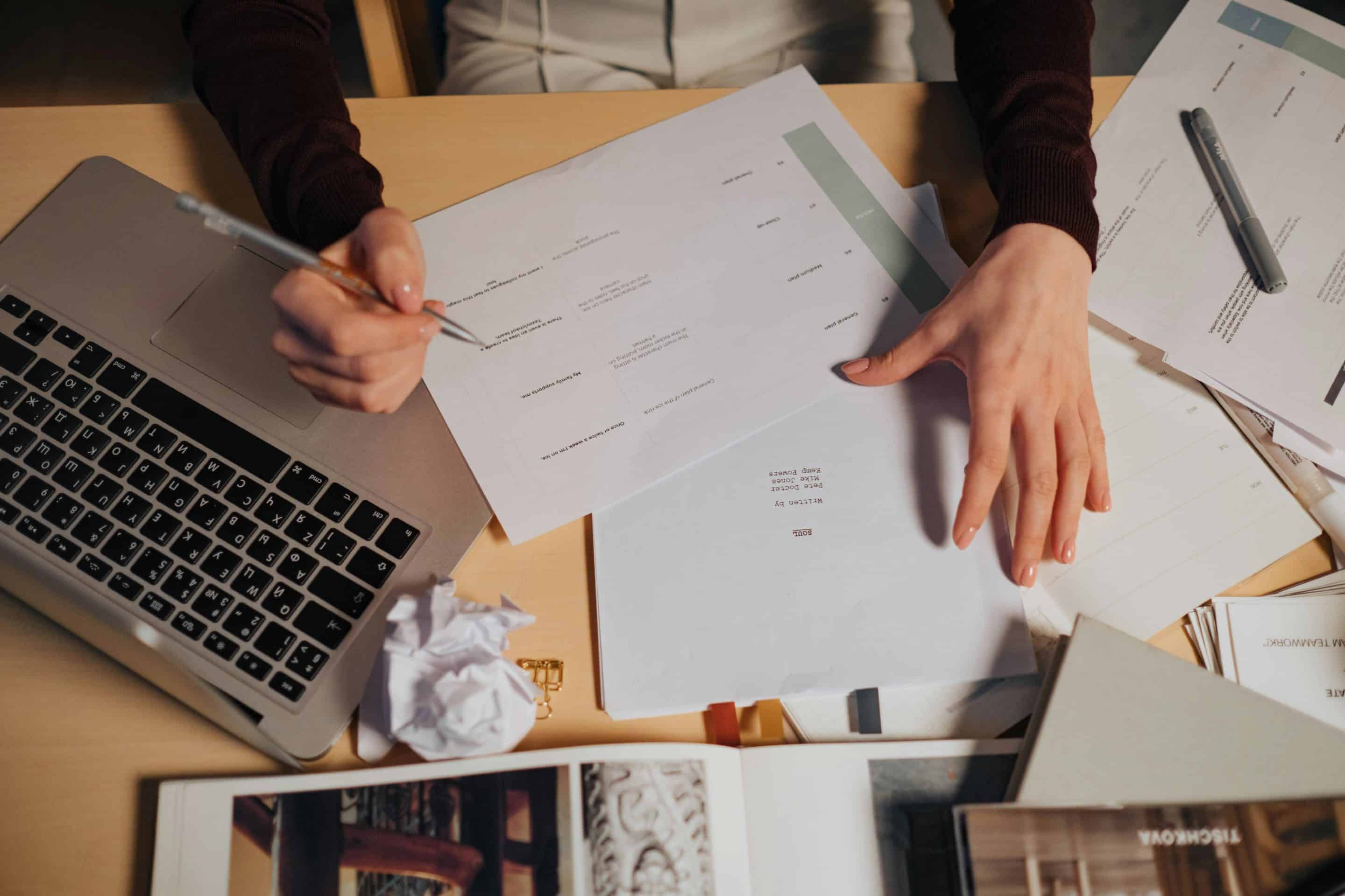View of person's hands while writing at a desk with scattered papers